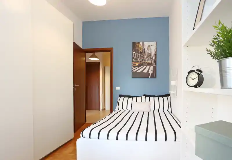 Bright, neatly staged single bedroom with a striped bed, decorative pillow and shelving. The room features a blue accent wall and wardrobe, ideal for a modern, compact rental.