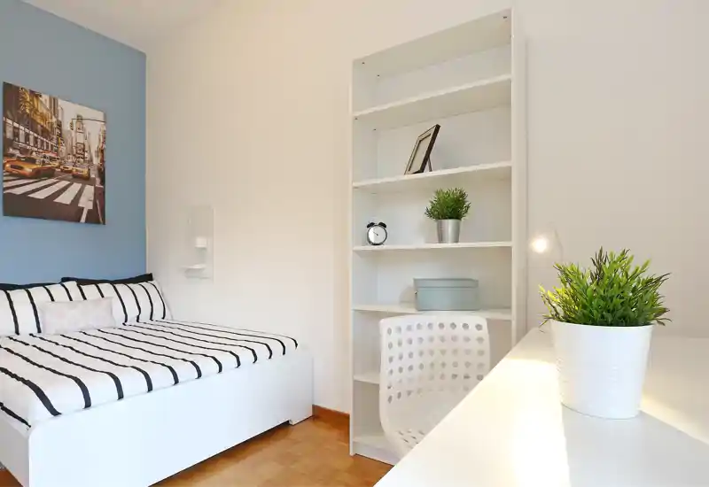 Bright, neatly styled bedroom with a white bed, striped linens, a storage shelf and a small desk with plants. The composition highlights calm, modern minimalism ideal for rental listings.