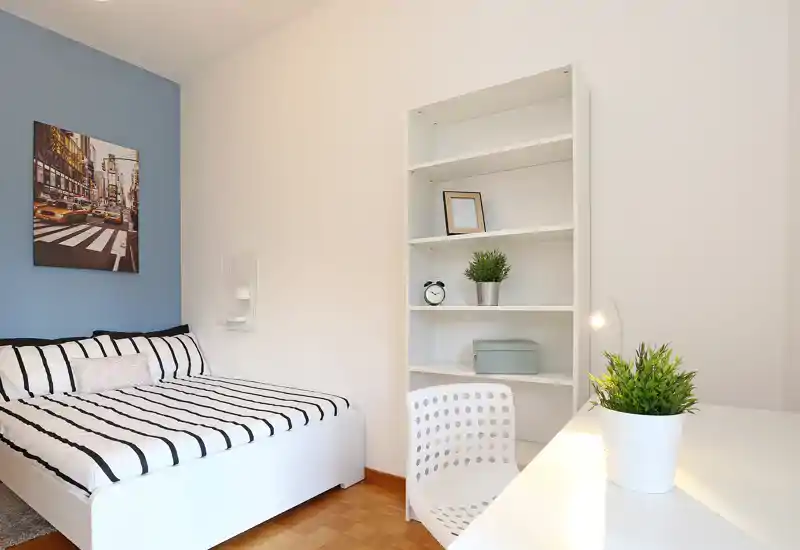 Bright, modern bedroom with a striped double bed, white shelving and a small desk area. The room features clean lines, decorative plants and soft accent colors for a welcoming feel.