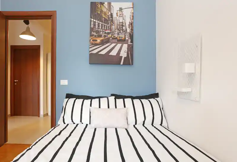 Bright, well-staged bedroom with a large striped bed, decorative pillow and framed city artwork above; clean lines and pleasant lighting make it inviting.