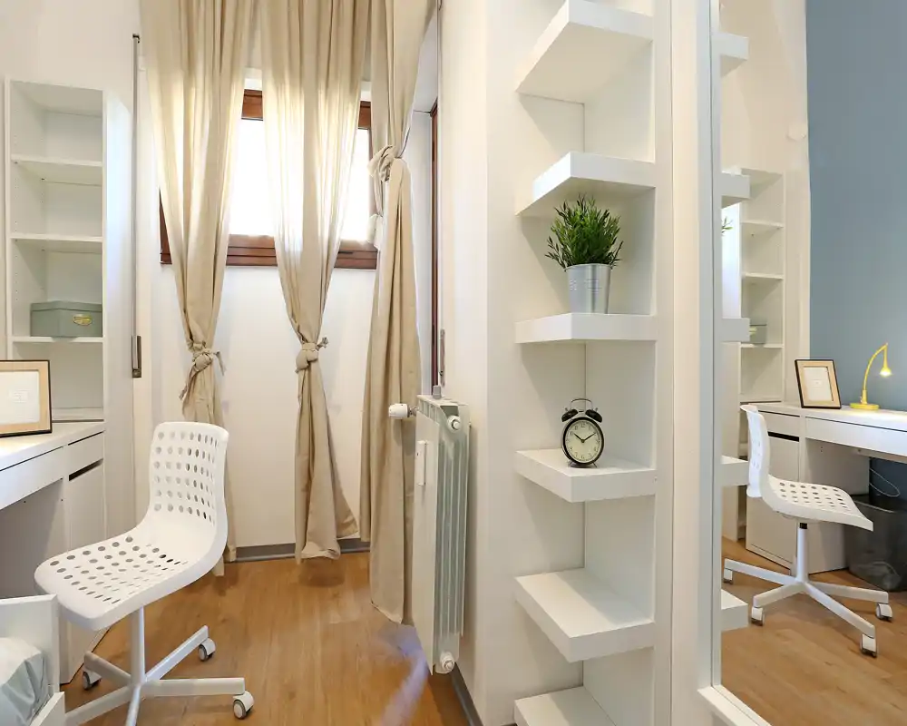 Espacio de estudio luminoso y compacto con escritorios y sillas blancas modernas, luz natural desde una ventana con cortinas y estanterías ordenadas – ideal para trabajar concentrado.