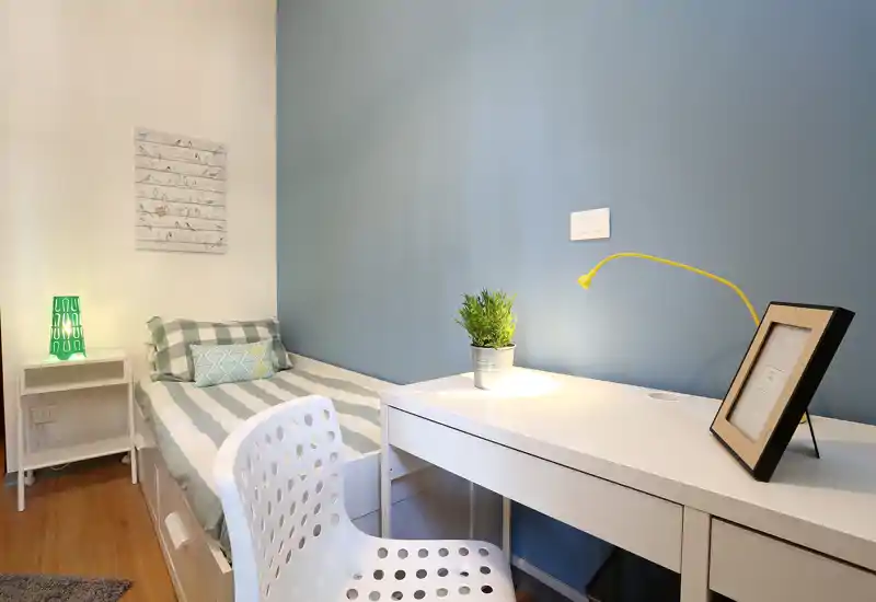 Dormitorio individual compacto y bien decorado con cama hecha, escritorio blanco, lámpara y pared azul tranquila — luminoso y acogedor, ideal para estudiar o descansar.