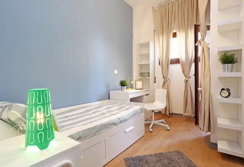 Dormitorio individual moderno y luminoso con cama blanca empotrada, escritorio y luz natural cálida desde la ventana con cortinas; decoración minimalista y suelo de madera que aportan confort.