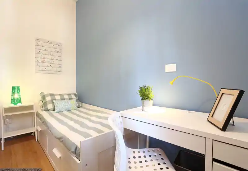 Dormitorio pequeño y luminoso con cama individual, mesita y escritorio blanco — pared azul y textiles acogedores que crean un rincón para dormir y trabajar.
