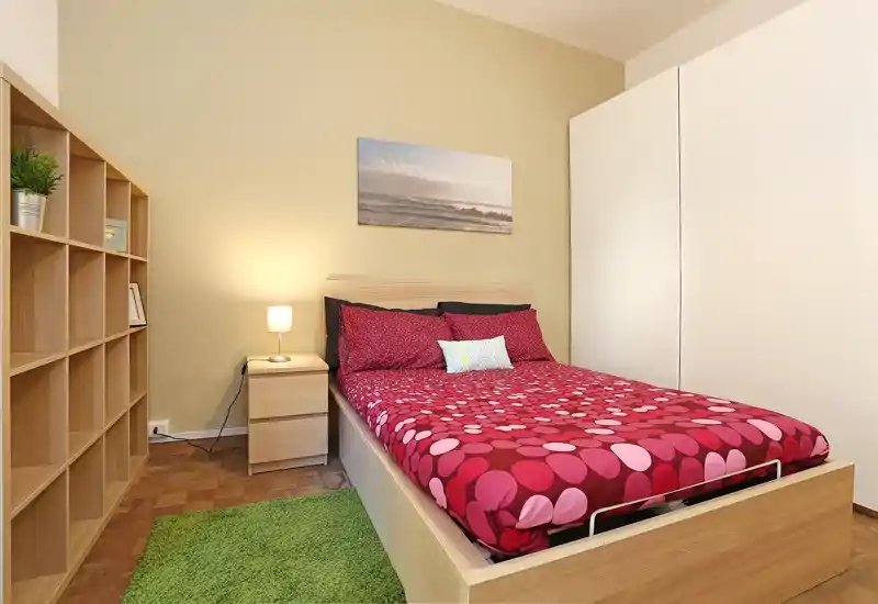 Dormitorio luminoso y bien decorado con cama doble con ropa de cama roja estampada, mesita con lámpara, estantería y gran armario — acogedor y listo para fotos.