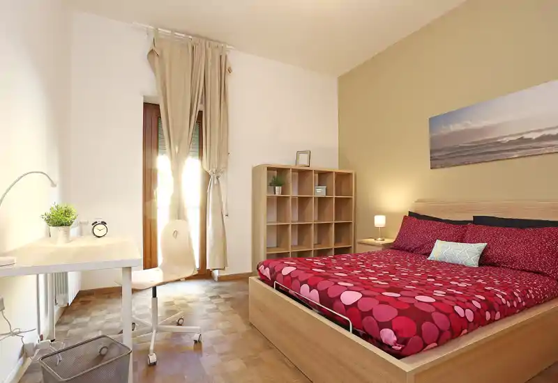 Dormitorio luminoso y moderno con cama de madera y ropa de cama roja de estampado, escritorio junto a la ventana y estantería abierta: limpio, bien iluminado y acogedor.