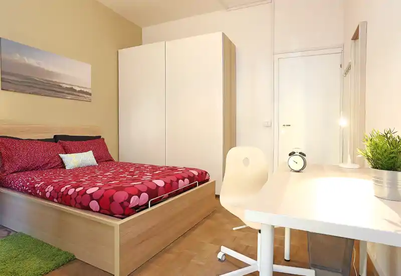 Dormitorio luminoso y bien presentado con cama de madera completa, armario y escritorio. La colcha roja estampada y la iluminación suave crean un espacio acogedor y ordenado, ideal para anuncios.