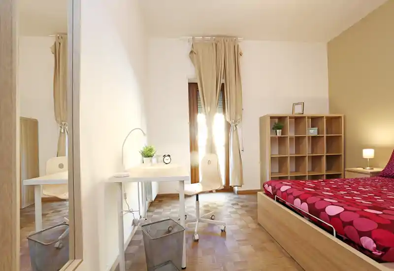 Camera da letto luminosa e ordinata con un letto matrimoniale in legno coperto da una trapunta rossa a motivo, una lampada da comodino e un mobile a scaffali. La luce naturale entra da una finestra alta con tende beige; un piccolo scrittoio bianco con sedia offre uno spazio studio.