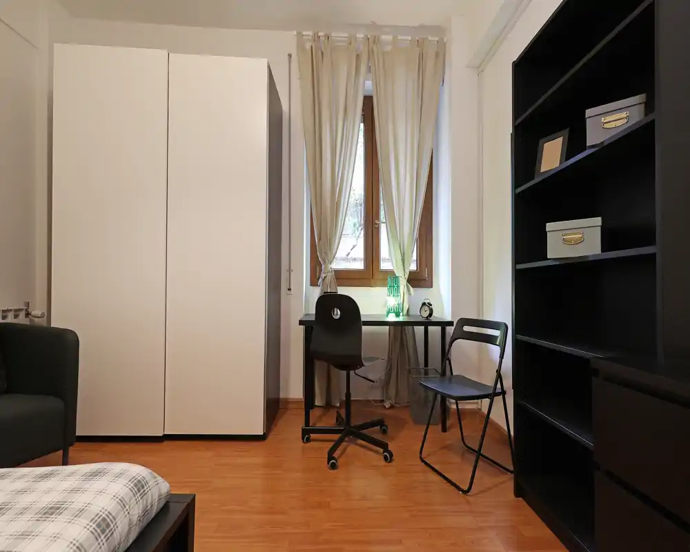 Angolo camera ben illuminato con letto visibile, armadio, scrivania vicino alla finestra e scaffalatura nera — stile pulito e moderno ideale per un annuncio.