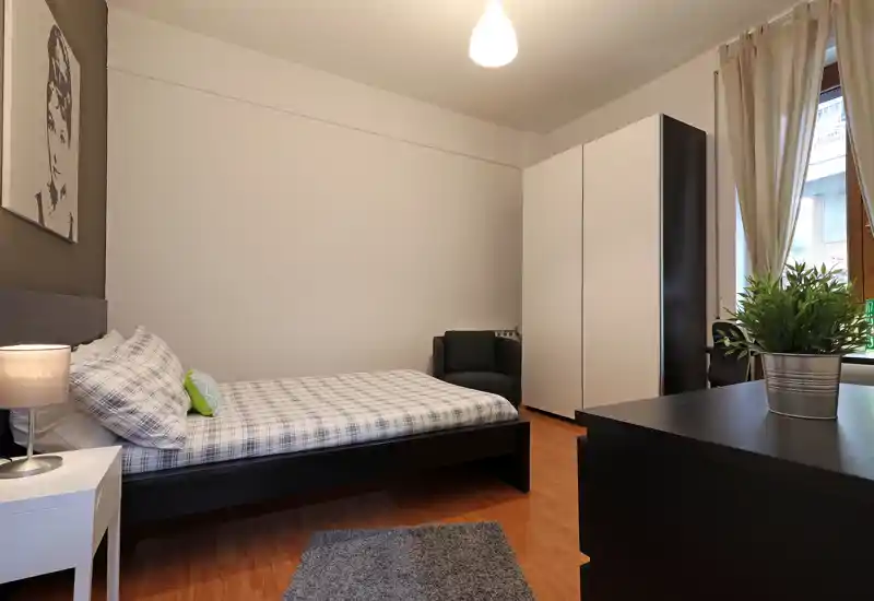 Dormitorio luminoso y bien presentado con cama doble, mesita de noche y armario. Suelo de madera y un pequeño rincón con sillón aportan una atmósfera moderna y acogedora.