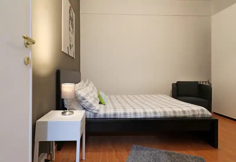 Dormitorio luminoso y minimalista con cama doble en estructura oscura, ropa de cama a cuadros, mesita con lámpara y un pequeño sillón en la esquina — líneas limpias y suelo de madera cálida.