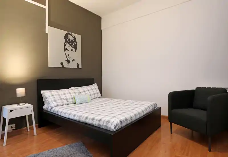 Dormitorio doble bien presentado con cama baja de estructura negra, ropa de cama a cuadros, mesita con lámpara y un sillón cómodo — moderno y acogedor.