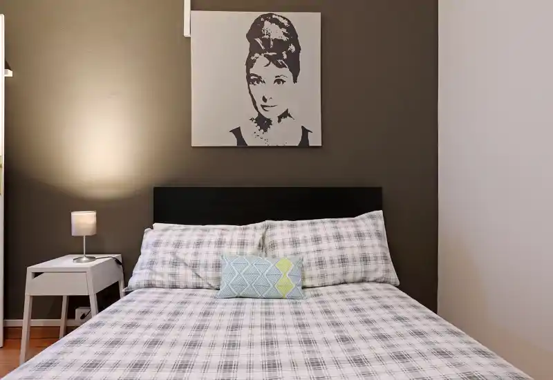Dormitorio luminoso y bien presentado con cama doble, ropa de cama estampada y mesita con lámpara. Tonos neutros y un cuadro en la pared crean un ambiente moderno y relajante.