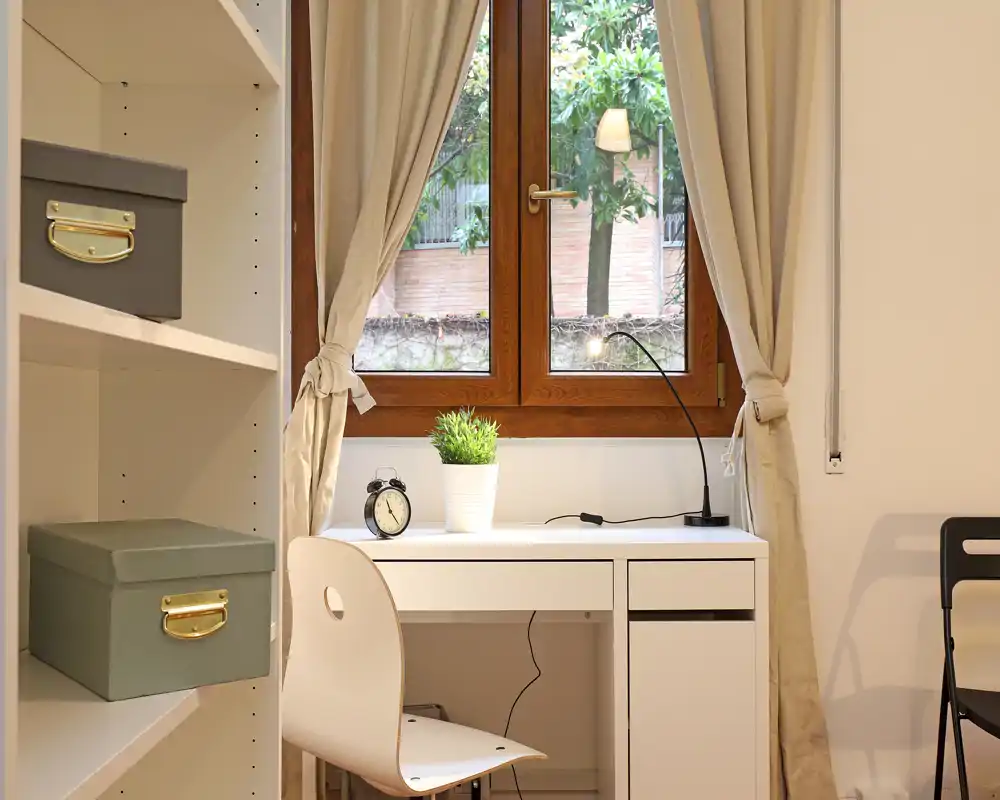 Angolo studio luminoso vicino alla finestra con scrivania bianca, sedia, lampada da tavolo e una pianta decorativa — perfetto per smart working o studio.