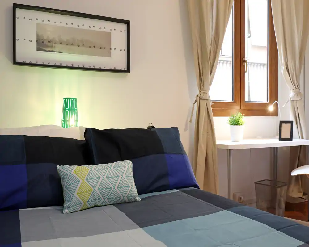 Camera da letto luminosa e ben arredata con piumone a motivi, cuscino decorativo e lampada da comodino; una piccola scrivania vicino alla finestra aggiunge funzionalità e luce naturale.