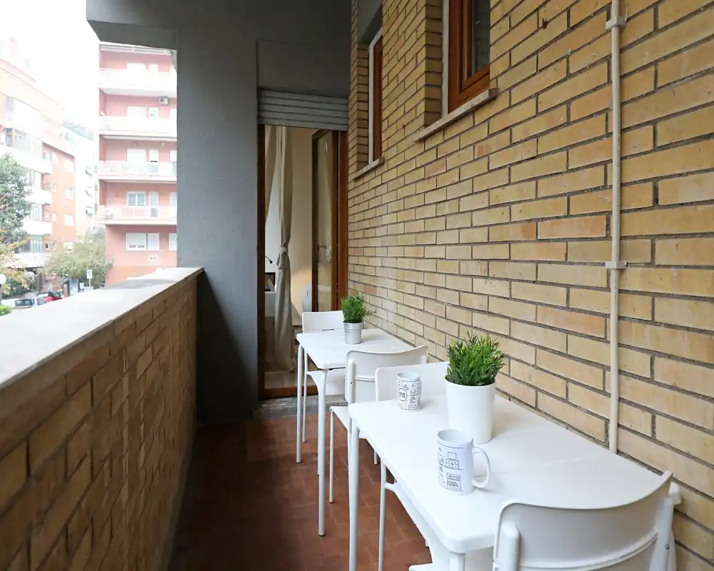 Balcone stretto e luminoso con parete in mattoni, due piccoli tavoli bianchi con sedie e piante in vaso — ideale per il caffè mattutino con vista sulla strada.