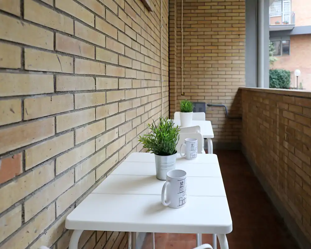 Balcone stretto e soleggiato con tavolini bianchi, piante in vaso e tazze — un angolo esterno accogliente per il caffè del mattino.