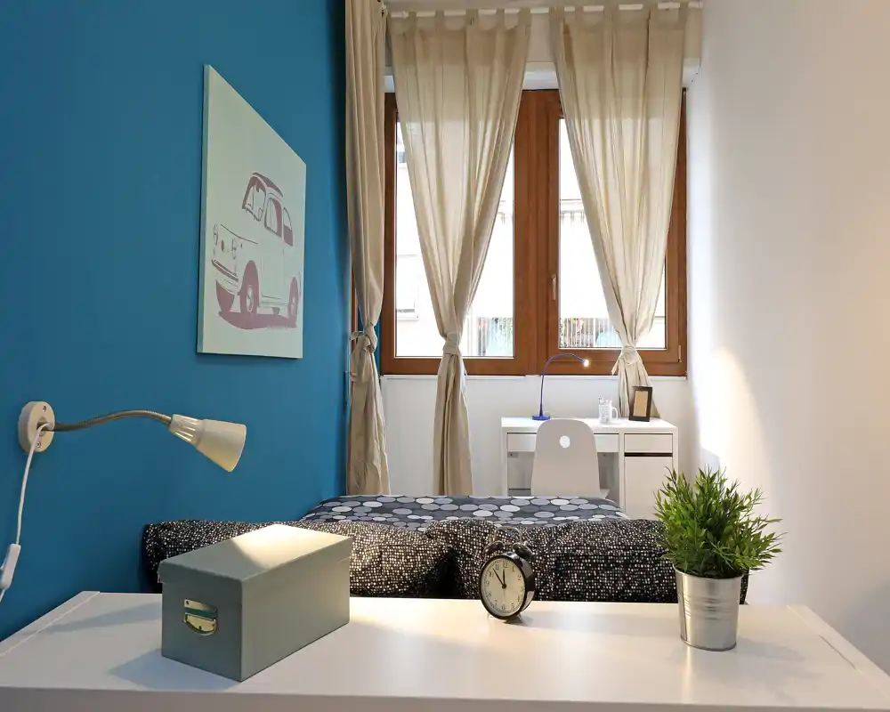 Camera singola moderna e luminosa con parete blu a contrasto, letto ordinato e una scrivania vicino alla finestra — perfetta per studenti o affitti brevi.