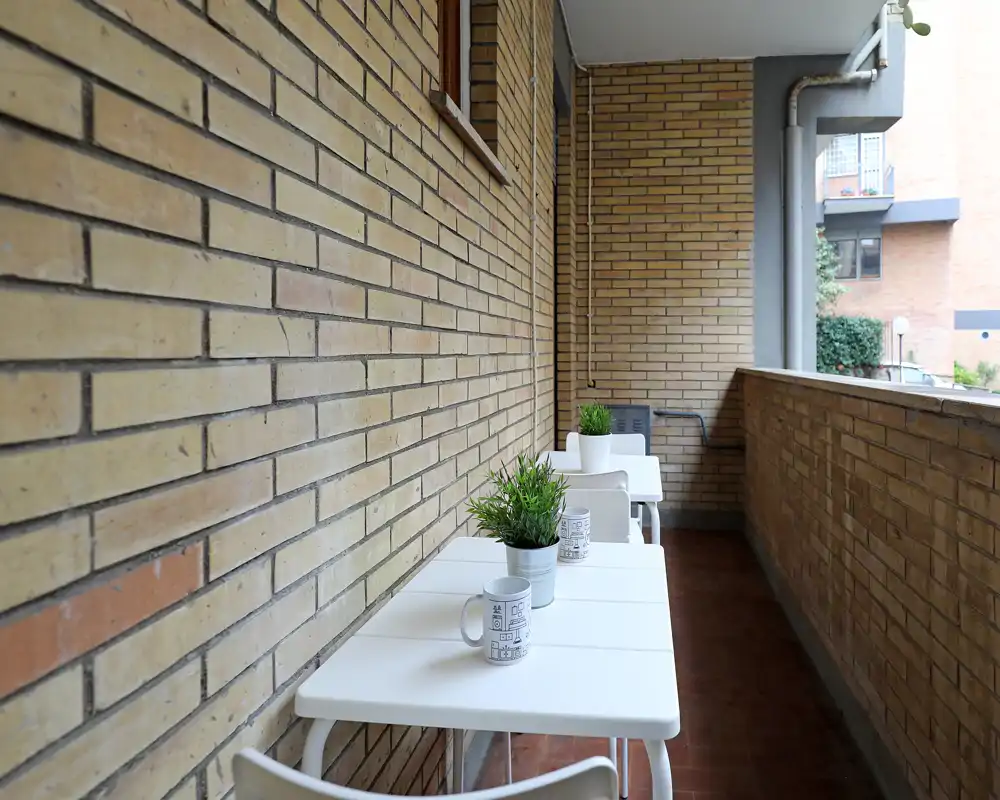 Balcone stretto e soleggiato con tavolini bianchi, piante in vaso e muro in mattoni — un angolo esterno accogliente per un caffè.
