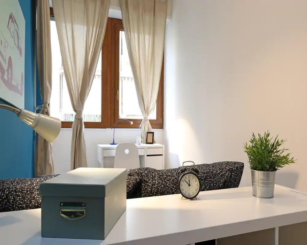 Angolo camera luminoso e minimal con letto visibile, scrivania sotto la finestra e luce naturale morbida—accogliente e moderno.