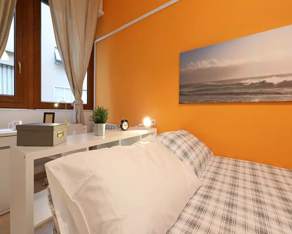 Camera da letto luminosa e accogliente con letto a quadri, parete arancione calda e mensola con lampada e sveglia — ideale per affitti brevi.