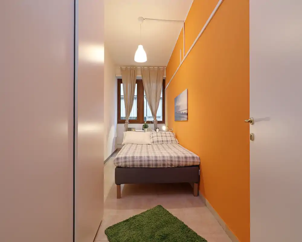 Camera da letto luminosa e compatta con letto matrimoniale centrato su una parete arancione, tende leggere e una lampada sul comodino che creano un ambiente accogliente.