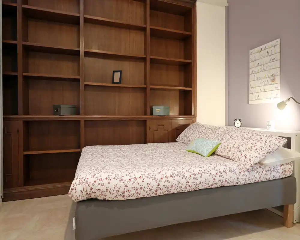 Camera da letto luminosa e accogliente con un letto matrimoniale a fiori posizionato davanti a un grande scaffale in legno incassato; comodino ordinato e luce soffusa per un'atmosfera calda.