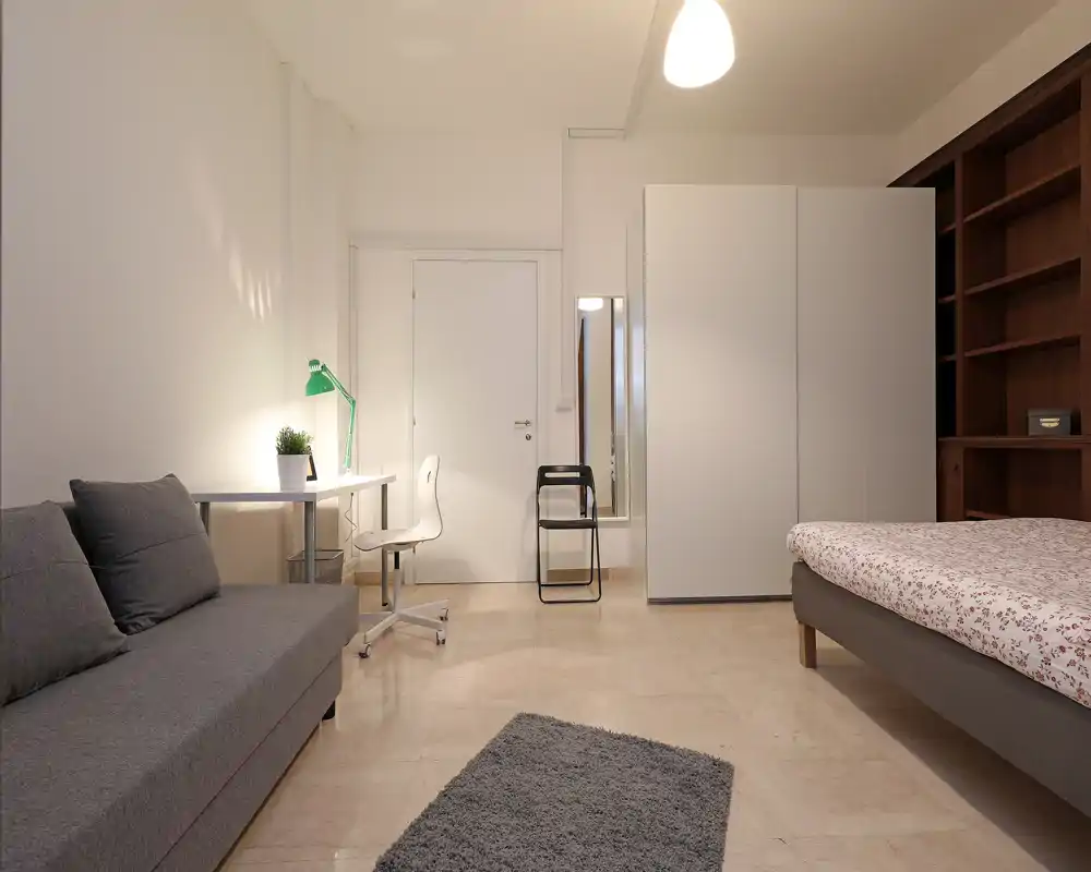 Camera da letto luminosa e ben arredata con letto matrimoniale visibile, divano, scrivania e scaffalature; toni neutri e illuminazione morbida creano un ambiente accogliente e adatto agli annunci.