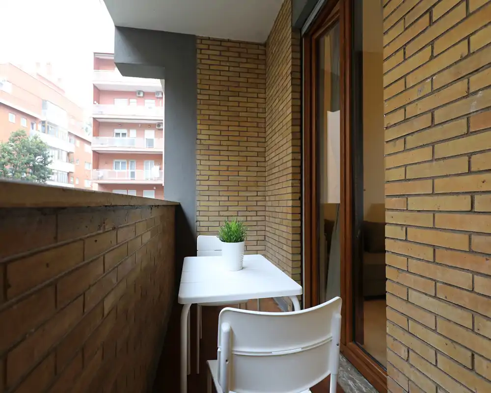 Accogliente balcone cittadino con un piccolo tavolo bianco, due sedie e una pianta in vaso — ideale per il caffè del mattino. Lo spazio si affaccia sul cortile condominiale e comunica con il soggiorno tramite una porta finestra scorrevole.