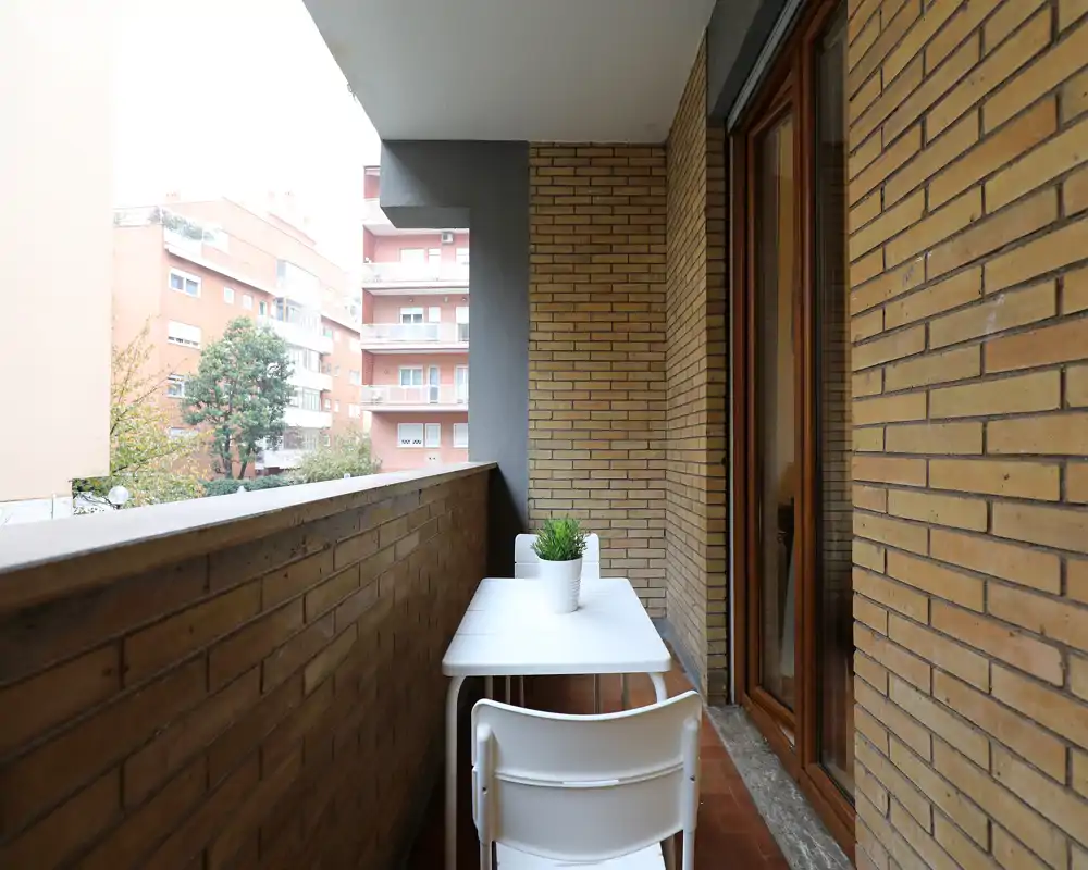 Balcone stretto e luminoso con piccolo tavolo bianco e due sedie, pareti in mattoni e una piantina in vaso — perfetto per il caffè mattutino.