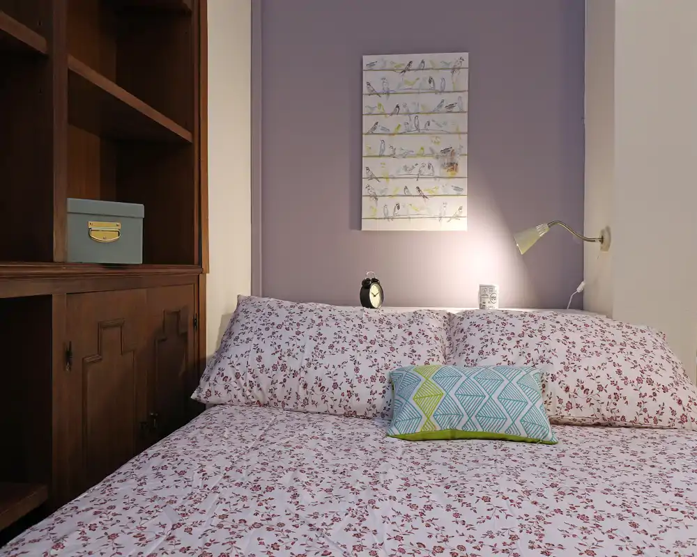 Camera da letto luminosa e accogliente con biancheria floreale, cuscino decorativo e lampada da comodino su una parete lilla; scaffalatura in legno integrata per spazio e calore.