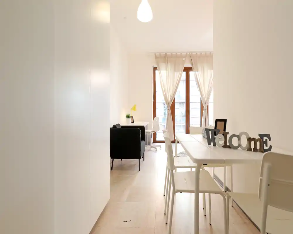 Zona giorno luminosa e minimal con un piccolo angolo salotto, una scrivania e un tavolo da pranzo vicino a grandi finestre con tende leggere.