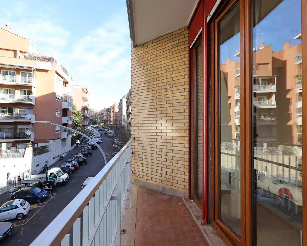 Balcone privato soleggiato con pavimento in cotto, affaccio su una strada residenziale e ampie porte scorrevoli in vetro che si aprono sull'appartamento.