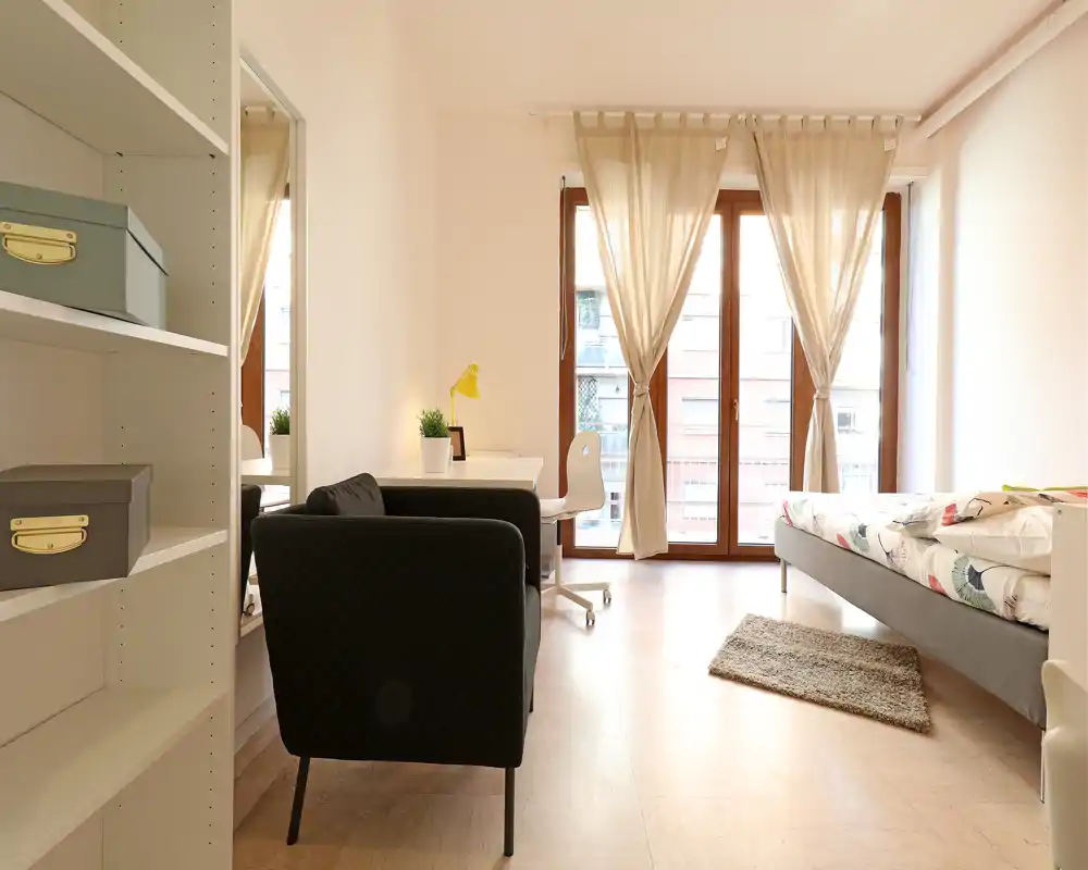 Camera da letto luminosa e moderna con grandi finestre a tutta altezza, un letto singolo con copripiumino decorato, una scrivania con sedia e una poltrona confortevole — perfetta per un appartamento cittadino accogliente.