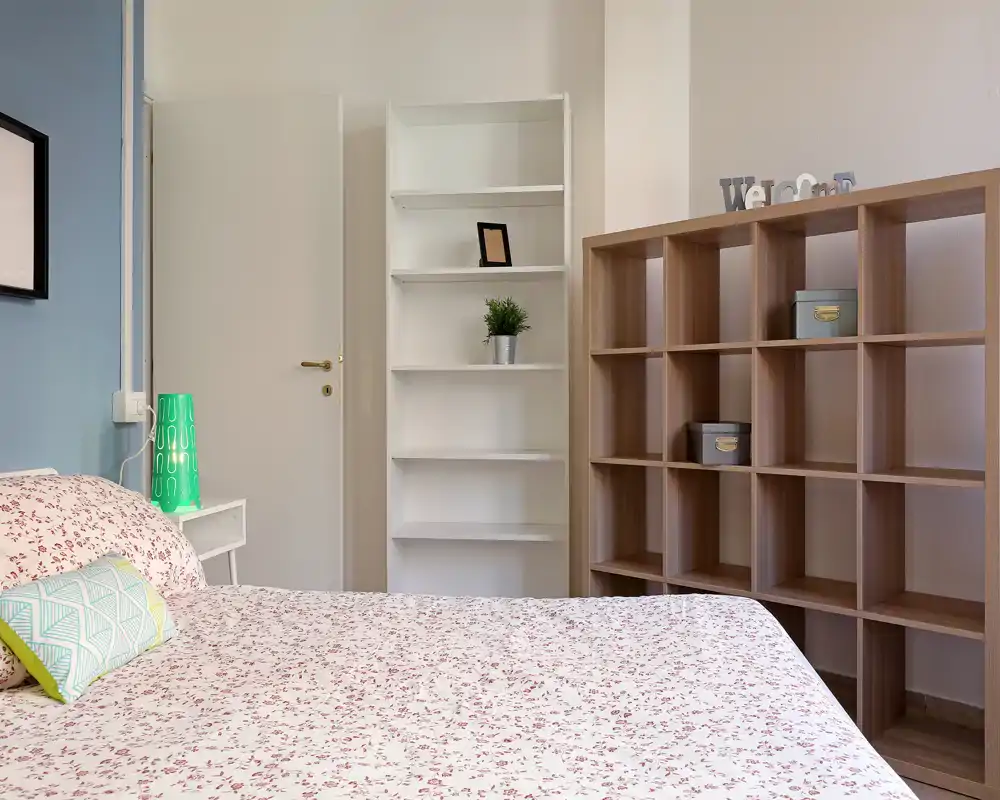 Camera da letto luminosa e accogliente con biancheria decorata, cuscini e scaffalature. L’inquadratura mette in evidenza il letto comodo, il comodino con lampada e lo spazio contenitivo ordinato.