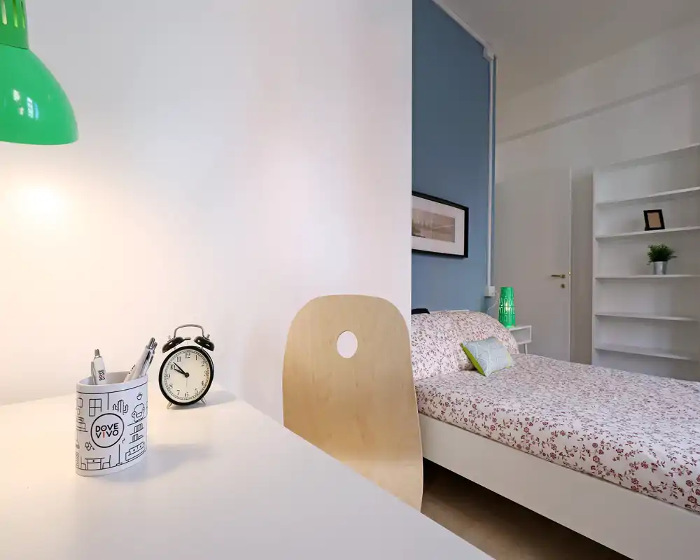 Camera da letto luminosa e ben curata con letto matrimoniale a fantasia, lampada da comodino e area scrivania minimal — perfetta per affitti brevi o studenti.