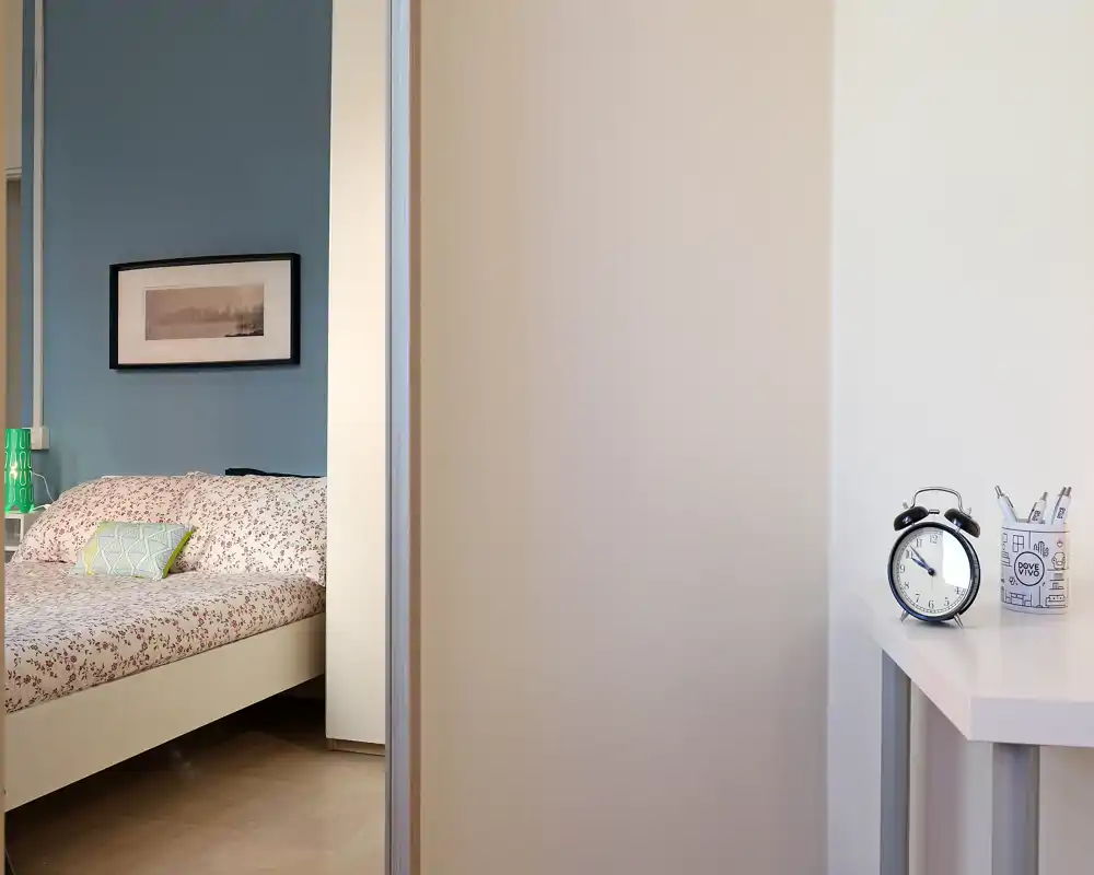 Angolo di camera da letto luminoso e curato, visto attraverso una porta, con letto a fantasia, cuscini decorativi e una lampada sul comodino che creano un’atmosfera accogliente.