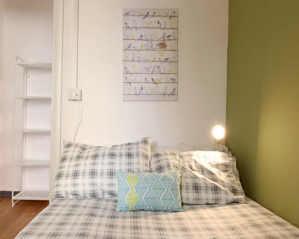 Camera da letto luminosa e ordinata con copriletto a quadri, cuscino decorativo e lampada da comodino calda: pulita, accogliente e ben allestita per foto d'affitto.