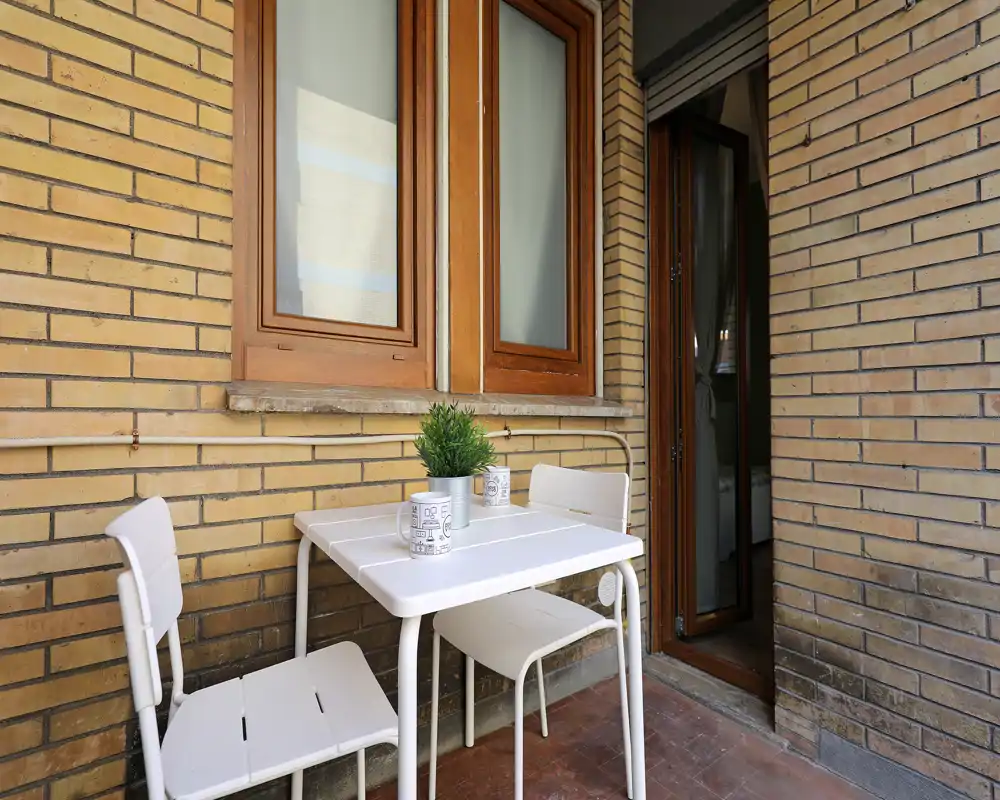 Piccolo balcone soleggiato con tavolino e sedie bianche in stile bistro appoggiati a una parete in mattoni — ideale per il caffè mattutino.