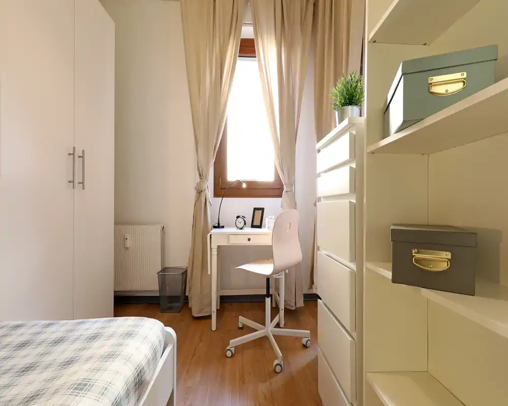 Camera da letto luminosa e ordinata con letto singolo, scrittoio davanti alla finestra e mobili contenitori ben organizzati; pavimento in legno e tende morbide creano un'atmosfera accogliente.