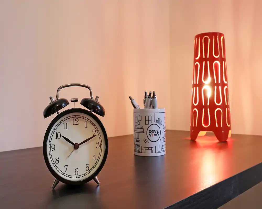 Primo piano di un piano d'appoggio con una sveglia vintage, un portapenne e una lampada rossa decorativa che diffonde una luce calda—ideale per mostrare dettagli e illuminazione.