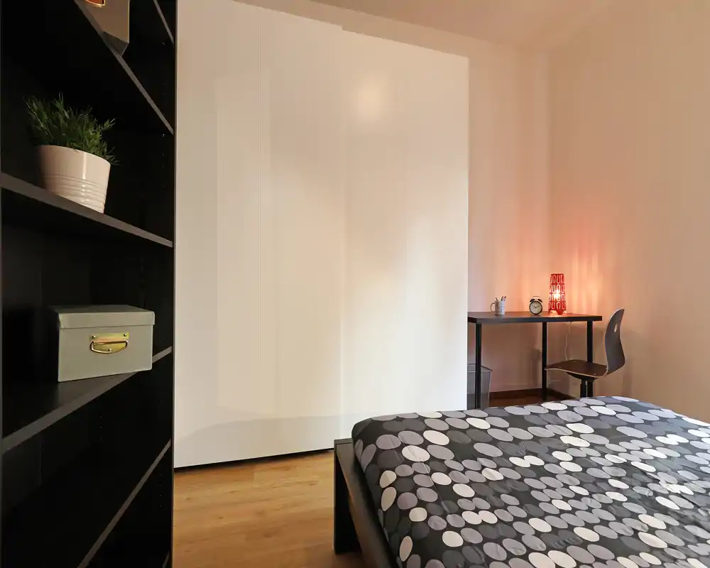 Camera da letto moderna e luminosa con copriletto a fantasia, ampio armadio bianco, mensole e una piccola scrivania con lampada: spazio tranquillo e ben allestito perfetto per annunci.