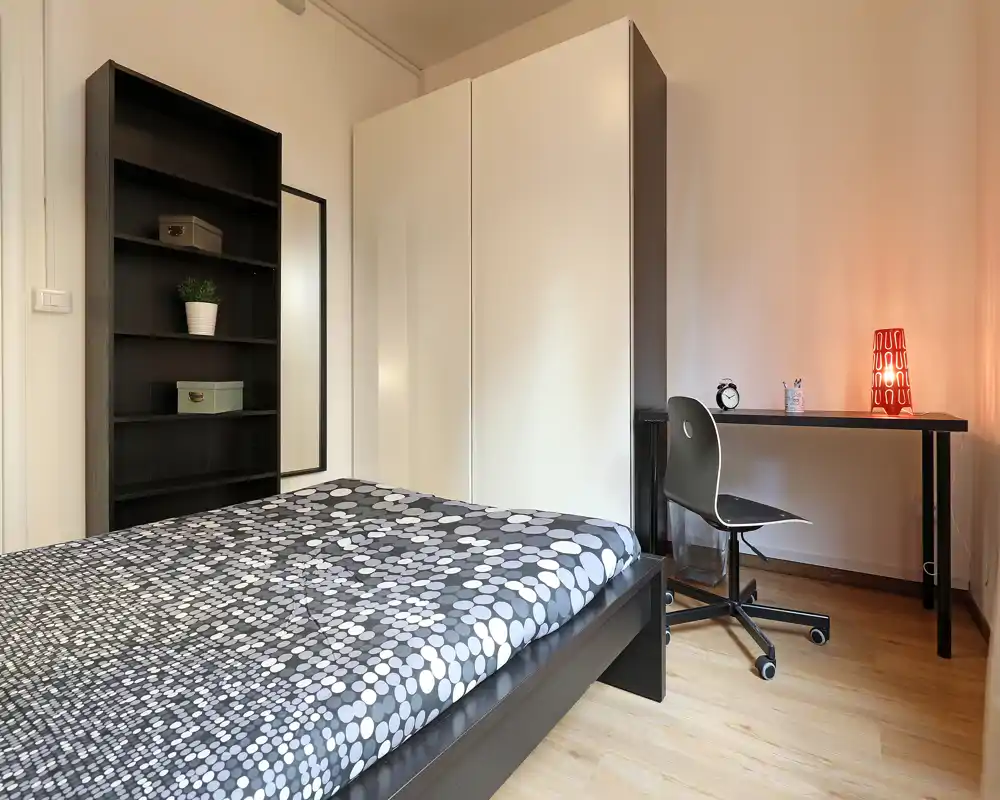 Camera da letto moderna e luminosa con un ampio letto in primo piano, armadio bianco, libreria e una scrivania con lampada — ambiente confortevole e ben illuminato, ideale per una o due persone.