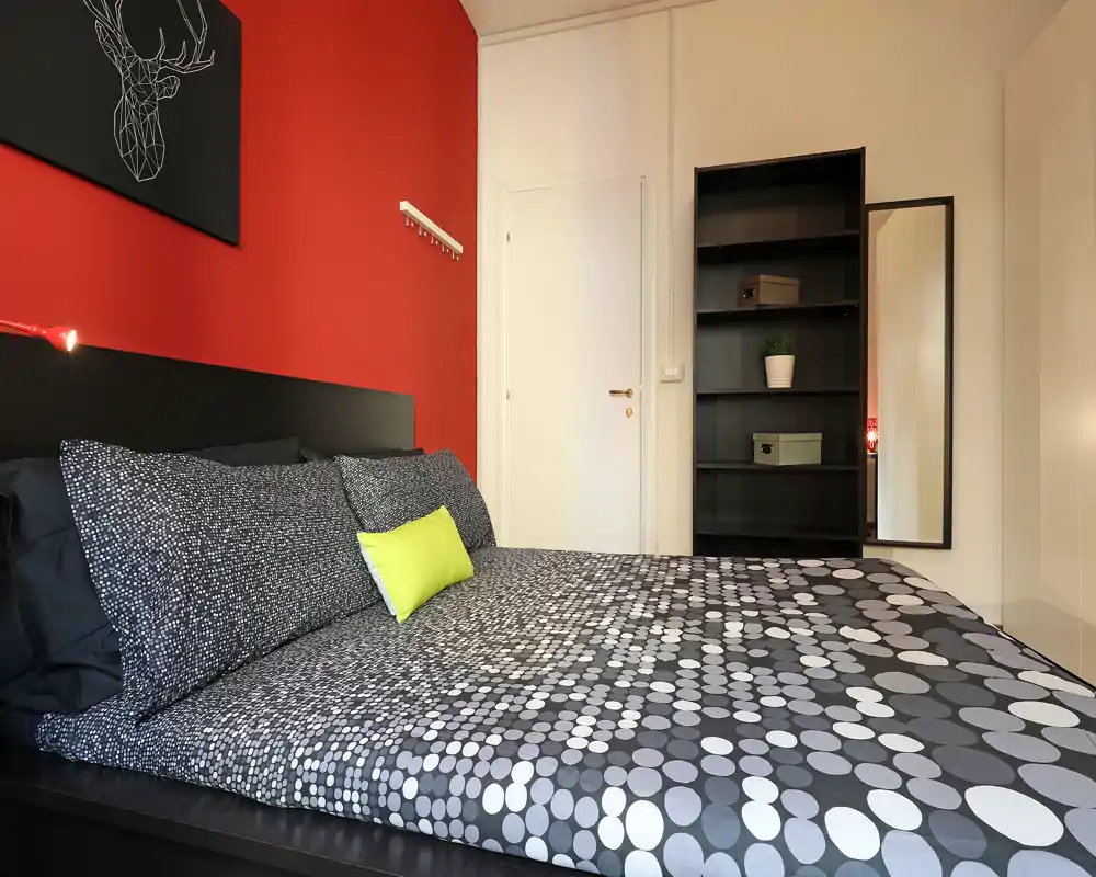 Camera da letto elegante con letto ampio dal copriletto a pois, parete d'accento rossa e scaffalatura ordinata — ambiente luminoso e moderno adatto ad affitti.