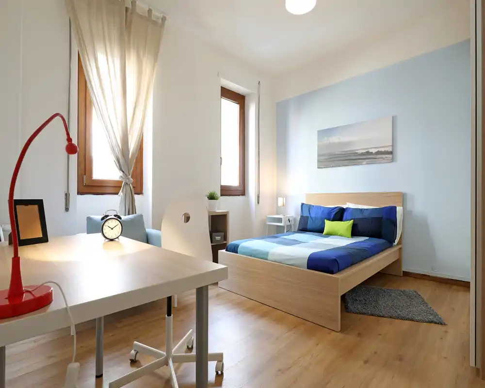 Camera da letto luminosa e moderna con letto in legno basso con biancheria blu, comodino e una piccola scrivania vicino a due finestre che lasciano entrare luce naturale.