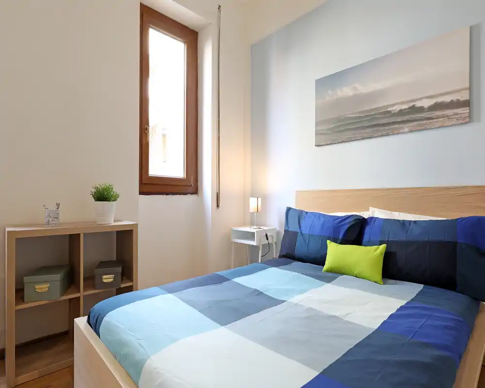 Camera da letto luminosa e ordinata con letto matrimoniale con copriletto blu, comodino con lampada e una stampa marina sopra la testata. La luce naturale entra dalla finestra, creando un ambiente fresco e accogliente ideale per gli annunci.