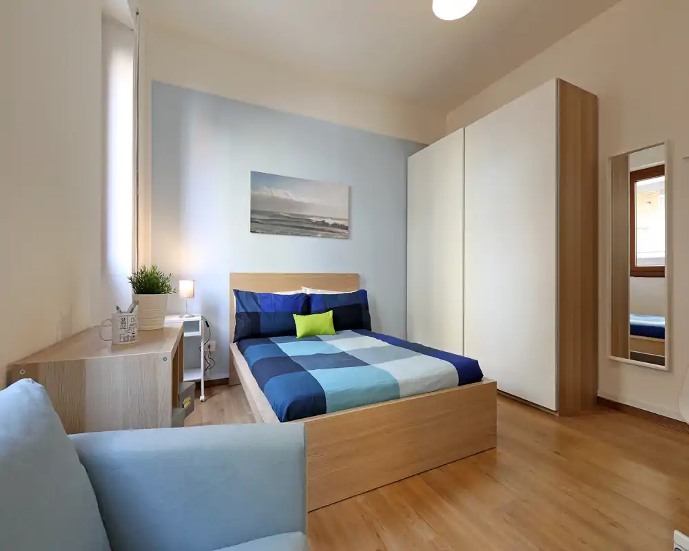Camera da letto moderna e luminosa con letto in legno e copriletto blu a quadri, ampia armadiatura e zona salotto: perfetta per annunci in affitto che puntano su arredi sobri e luce naturale.