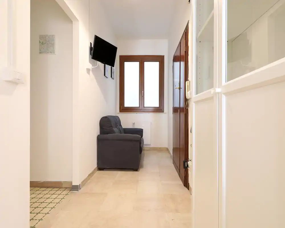 Soggiorno stretto con un piccolo divano due posti, TV a parete e una finestra che dona luce naturale — ambiente compatto e arredamento essenziale.