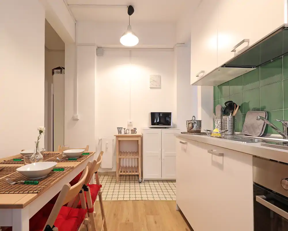 Cucina compatta e luminosa con mobili bianchi, schienale in piastrelle verdi, lavello integrato e un piccolo tavolo da pranzo per quattro — ambiente accogliente e ben arredato per i pasti quotidiani.