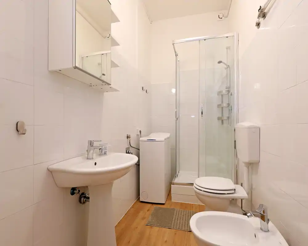 Bagno pulito e compatto con lavabo a colonna, WC, bidet, lavatrice e doccia con box in vetro. Piastrelle bianche e pavimento effetto legno donano un ambiente luminoso e funzionale.